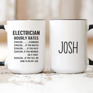 Peut inclure: Deux tasses à café blanches avec des poignées noires. Une tasse porte le texte "ELECTRICIAN HOURLY RATES" et une liste de tarifs. L'autre tasse porte le nom "JOSH".