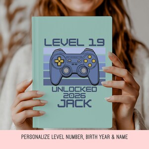 May include: A light green notebook with the text "LEVEL 19 UNLOCKED 2026 JACK" and a blue video game controller graphic. The notebook is held by a person.