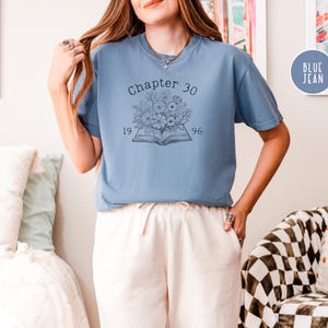 May include: A blue jean-colored t-shirt with the text "Chapter 30" above a floral and book graphic. The numbers "1996" and "19" are below the book. The shirt is a comfortable, casual style.