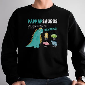 Peut inclure: Sweat-shirt noir avec un motif de dinosaure vert et le texte "PAPPAPSAURUS Comme un Pap Pap ordinaire, mais plus RAWESOME !" avec les noms "MILLY", "FINN", "VIOLET" et "MILES" en dessous.