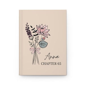 May include: A beige journal with a floral illustration and the text "Anna CHAPTER 65." The illustration features a bouquet of flowers in pink, purple, and green, tied with a pink ribbon. The journal has a clean, minimalist design.