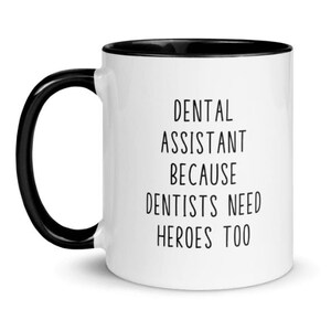 May include: White ceramic coffee mug with a black rim and handle. The mug has black text that reads "Dental Assistant Because Dentists Need Heroes Too."