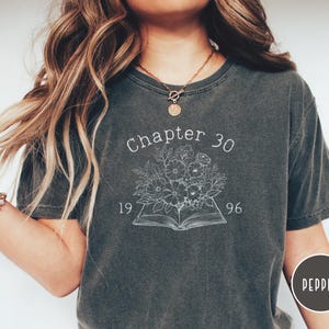 May include: Dark gray t-shirt with the text "Chapter 30 1996" above a floral design over an open book. The shirt is a crew neck style and has a slightly faded appearance. The shirt is a comfortable, casual wear.