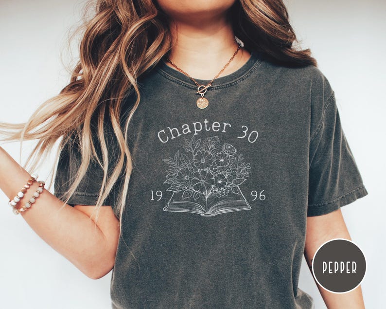 May include: Dark gray t-shirt with the text "Chapter 30 1996" above a floral design over an open book. The shirt is a crew neck style and has a slightly faded appearance. The shirt is a comfortable, casual wear.