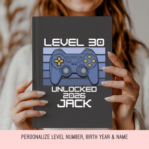 May include: A dark gray notebook featuring a blue video game controller graphic with the text "LEVEL 30 UNLOCKED 2026 JACK." The notebook has a personalized design, perfect for gamers celebrating a milestone birthday.