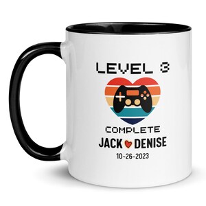 May include: White ceramic mug with a black handle and black rim. The mug has a graphic of a video game controller inside a heart with a retro sunset background. The text "LEVEL 3 COMPLETE JACK ♥ DENISE 10-26-2023" is printed below the graphic.