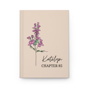 May include: A beige journal with a floral illustration and the name "Katelyn" and "CHAPTER 85" in black script. The floral illustration features pink flowers and green leaves.