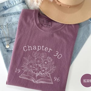 May include: Berry-colored t-shirt with the text "Chapter 30" and a floral design. The shirt is laid on top of light blue jeans, with a gold necklace and a tan hat in the background. The shirt also has the numbers "1996" on it.