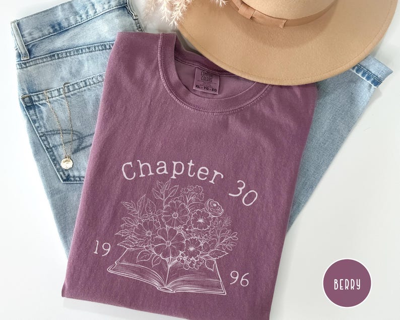 May include: Berry-colored t-shirt with the text "Chapter 30" and a floral design. The shirt is laid on top of light blue jeans, with a gold necklace and a tan hat in the background. The shirt also has the numbers "1996" on it.