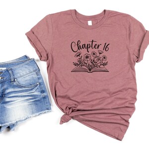 May include: A mauve t-shirt with the text "Chapter 16" and a black and white illustration of a book with flowers on top.