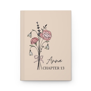May include: A beige journal with a floral illustration of a bouquet of pink flowers with a pink ribbon. The text "Anna Chapter 13" is printed below the illustration.