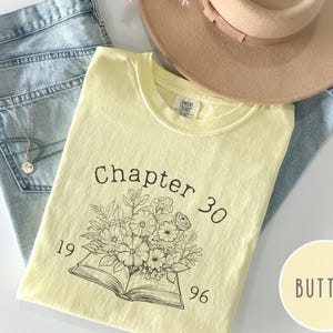 May include: A light yellow t-shirt with the text "Chapter 30 1996" and a floral design. The shirt is laid on top of blue jeans and a tan hat. A gold necklace is visible.
