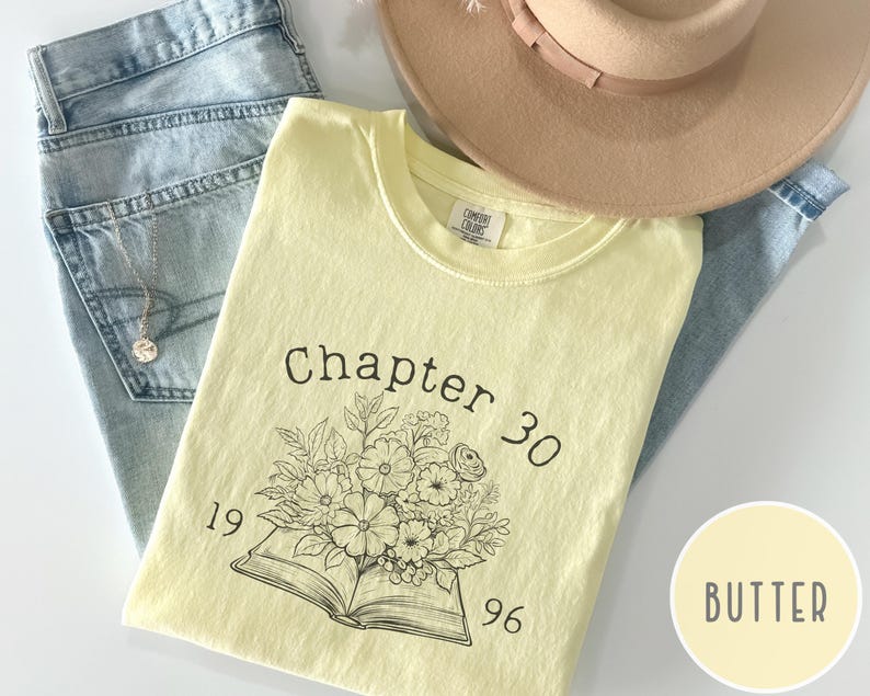 May include: A light yellow t-shirt with the text "Chapter 30 1996" and a floral design. The shirt is laid on top of blue jeans and a tan hat. A gold necklace is visible.