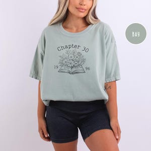 May include: A light green t-shirt with the text "Chapter 30" above an open book illustration with flowers and the numbers "1996". The shirt is paired with black shorts.