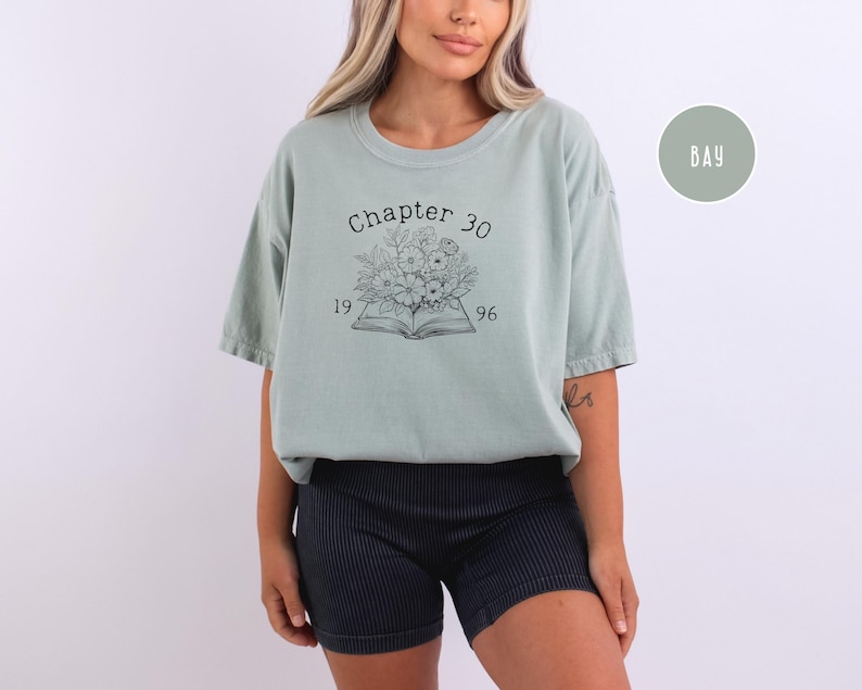 May include: A light green t-shirt with the text "Chapter 30" above an open book illustration with flowers and the numbers "1996". The shirt is paired with black shorts.