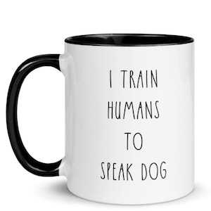 May include: White ceramic coffee mug with a black rim and handle. The mug has black text that reads "I train humans to speak dog."