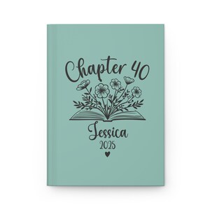 May include: A teal blue journal cover with a black illustration of an open book with flowers growing out of it. The text "Chapter 40" is above the book and the text "Jessica 2025" is below the book.
