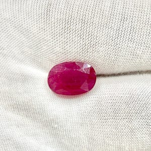 Op de afbeelding: Een gefacetteerde, ovaalvormige robijn edelsteen. De robijn heeft een diepe, levendige rode kleur en lijkt op een witte stoffen achtergrond te liggen. De facetten van de edelsteen vangen het licht op en creëren een sprankelend effect.