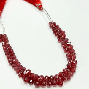 Puede incluir: Un collar de cuentas de piedras preciosas rojo oscuro facetadas. Las cuentas tienen forma de lágrima y están ensartadas, con una borla roja en la parte superior. Las cuentas probablemente se utilizan para la fabricación de joyas.