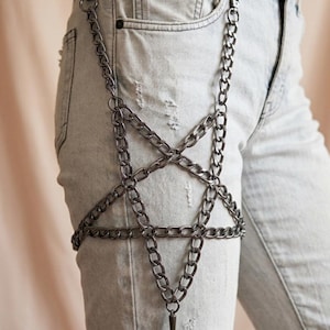 Puede incluir: Un arn&eacute;s de cadena plateada con dise&ntilde;o de pentagrama, usado sobre vaqueros de mezclilla de lavado claro. El arn&eacute;s presenta una serie de eslabones de cadena interconectados que forman una estrella en el muslo, con un colgante puntiagudo colgando del centro.