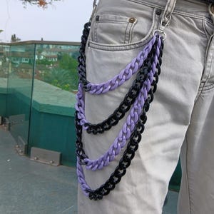 Può includere: Una persona indossa jeans grigi chiari con una cintura a catena a più strati. La catena è realizzata con maglie di plastica nera e viola.