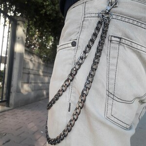 Op de afbeelding: Een zwarte ketting met een zilveren sluiting bevestigd aan een beige spijkerbroek.