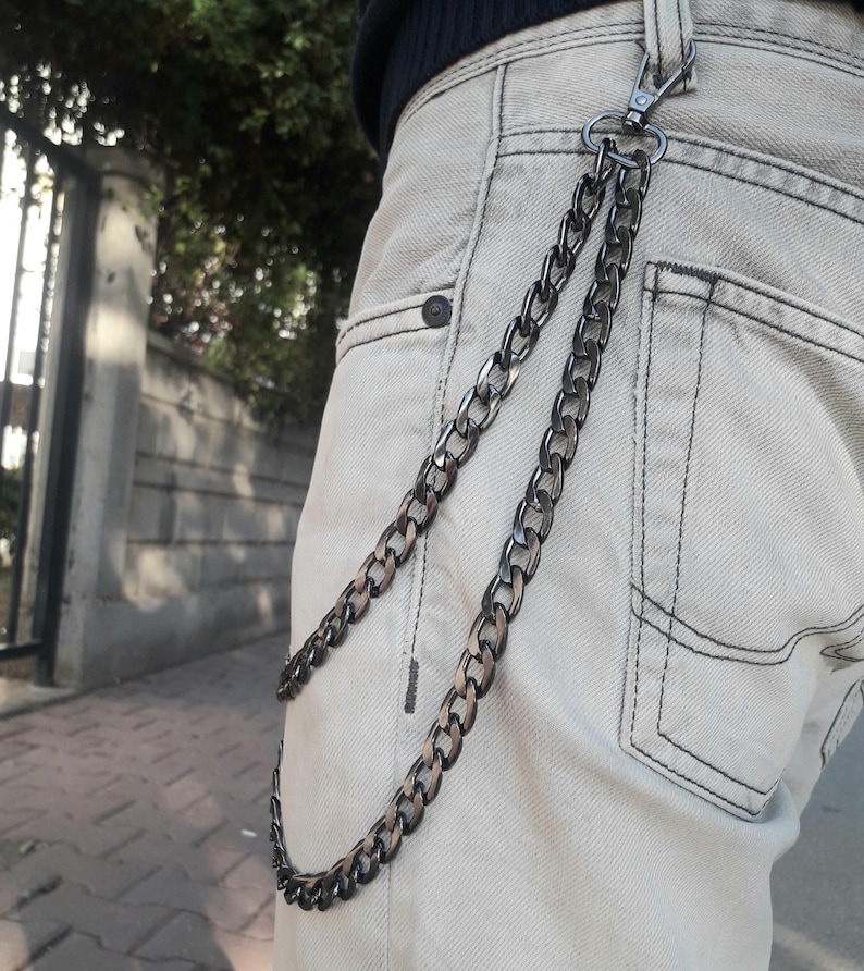 Op de afbeelding: Een zwarte ketting met een zilveren sluiting bevestigd aan een beige spijkerbroek.