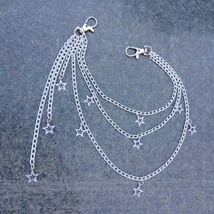 May include: Silver-colored chain accessory with multiple strands and star-shaped charms. The accessory features two clasp closures and is designed to be attached to clothing or accessories. The chains are layered, with stars dangling at varying lengths.