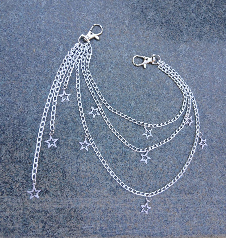 May include: Silver-colored chain accessory with multiple strands and star-shaped charms. The accessory features two clasp closures and is designed to be attached to clothing or accessories. The chains are layered, with stars dangling at varying lengths.