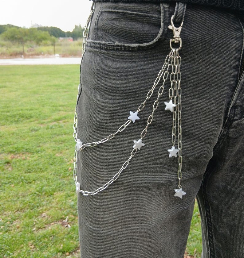 K&ouml;nnte beinhalten: Silberne Ketten-G&uuml;rtelkette mit Stern-Anh&auml;ngern. Die Kette ist an einem silbernen Verschluss befestigt und h&auml;ngt an der Vorderseite einer dunkelgrauen Jeans herunter. Die Stern-Anh&auml;nger sind wei&szlig;.