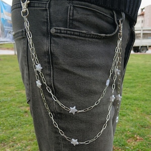K&ouml;nnte beinhalten: Silberkette mit sternf&ouml;rmigen Anh&auml;ngern, die an einer dunkelgrauen Jeans befestigt sind. Die Kette ist mit der G&uuml;rtelschlaufe und der Tasche der Jeans verbunden. Die Kette h&auml;ngt herunter.