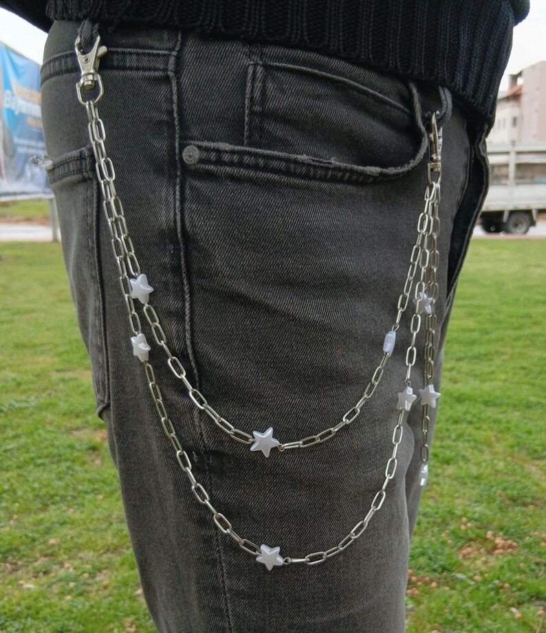 K&ouml;nnte beinhalten: Silberkette mit sternf&ouml;rmigen Anh&auml;ngern, die an einer dunkelgrauen Jeans befestigt sind. Die Kette ist mit der G&uuml;rtelschlaufe und der Tasche der Jeans verbunden. Die Kette h&auml;ngt herunter.