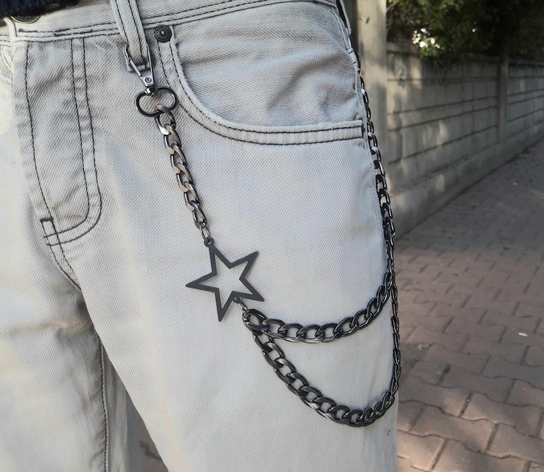 Op de afbeelding: Een zwarte ketting met een stervormige hanger eraan. De ketting hangt aan een riemlus aan een lichtgrijze spijkerbroek.