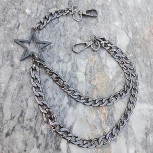 Op de afbeelding: Een zwarte metalen ketting met een stervormige hanger. De ketting heeft twee verschillende maten schakels en is bevestigd aan twee draaibare sluitingen.