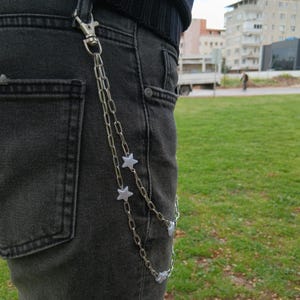 K&ouml;nnte beinhalten: Silberne Kette mit Sternanh&auml;ngern, befestigt an einer schwarzen Jeans. Die Kette wird mit einem Verschluss befestigt und h&auml;ngt in zwei Str&auml;ngen. Die Sternanh&auml;nger sind gleichm&auml;&szlig;ig &uuml;ber die Kette verteilt.