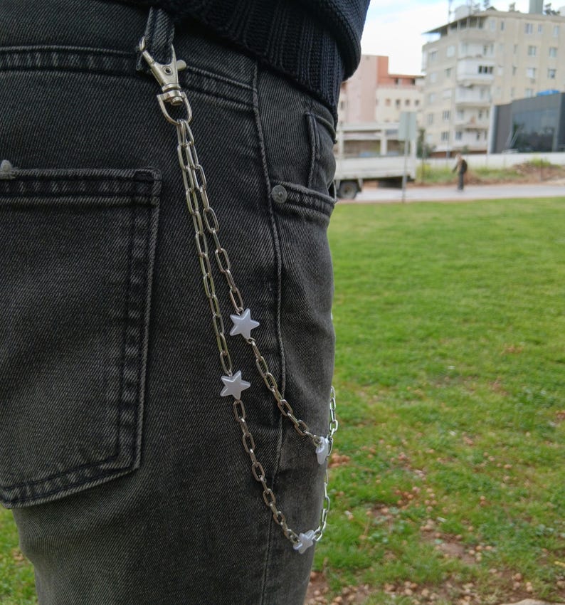 K&ouml;nnte beinhalten: Silberne Kette mit Sternanh&auml;ngern, befestigt an einer schwarzen Jeans. Die Kette wird mit einem Verschluss befestigt und h&auml;ngt in zwei Str&auml;ngen. Die Sternanh&auml;nger sind gleichm&auml;&szlig;ig &uuml;ber die Kette verteilt.