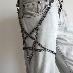 Puede incluir: Un accesorio de cadena plateada con un dise&ntilde;o de pentagrama, usado en el muslo. La cadena est&aacute; conectada con un colgante de punta. El accesorio se muestra en vaqueros de mezclilla de color claro.