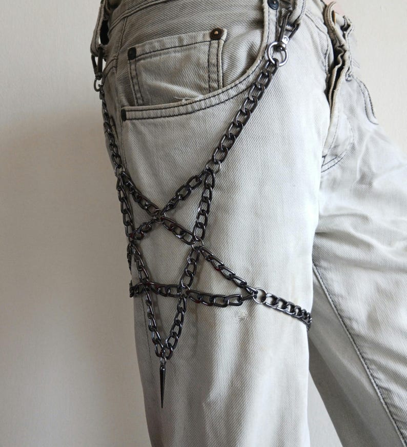 Puede incluir: Un accesorio de cadena plateada con un dise&ntilde;o de pentagrama, usado en el muslo. La cadena est&aacute; conectada con un colgante de punta. El accesorio se muestra en vaqueros de mezclilla de color claro.