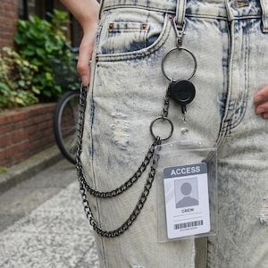 Könnte beinhalten: Ein silberfarbenes Ketten-Geldbörsen-Accessoire, das an hellblau gewaschenen Jeans befestigt ist. Die Kette verbindet sich mit einer einziehbaren Ausweisrolle und einem transparenten Ausweishalter mit dem Text "ACCESS" und "CREW".