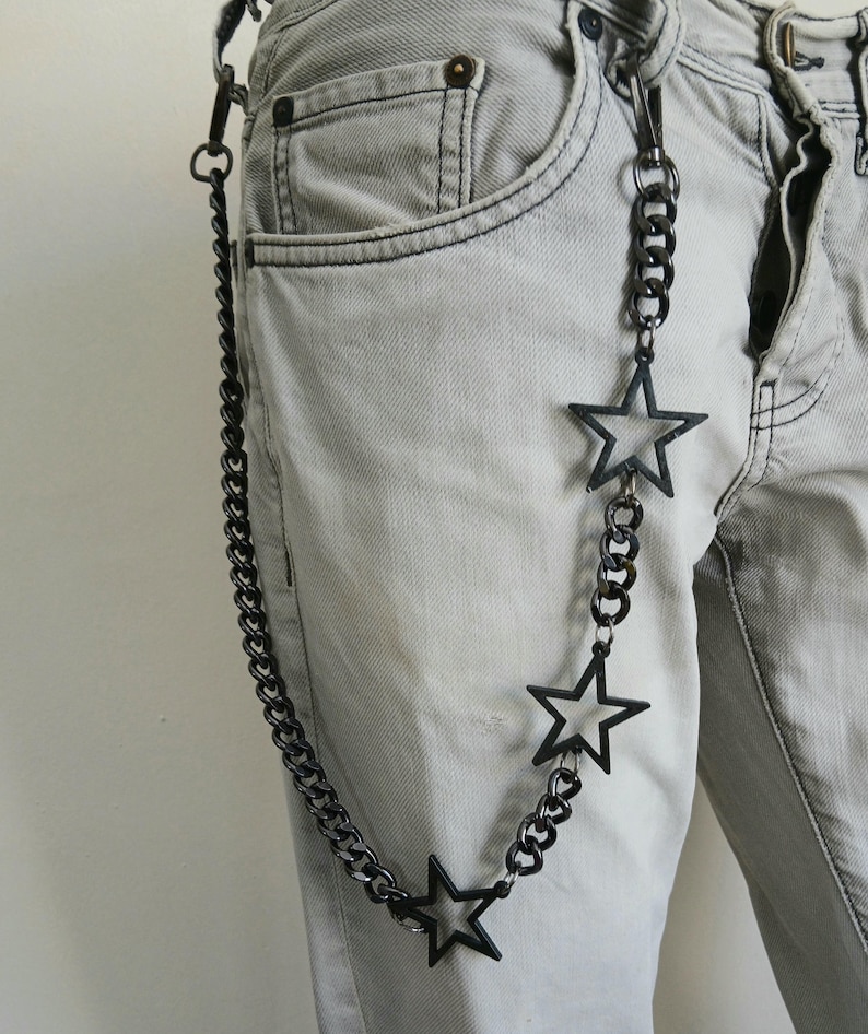 May include: A black chain with three star-shaped charms hangs from a belt loop on a pair of light gray jeans.