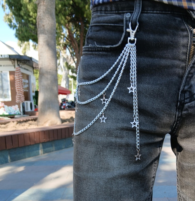 May include: Silver chain belt with star charms attached to a silver clasp. The chain hangs down in multiple strands, with star-shaped charms dangling from the chains. The belt is attached to a pair of dark gray denim jeans.