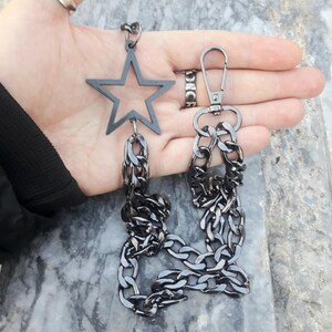 Op de afbeelding: Een zwarte metalen ketting met een stervormige hanger en een karabijnhaak. De ketting is dik en heeft een robuust ontwerp.