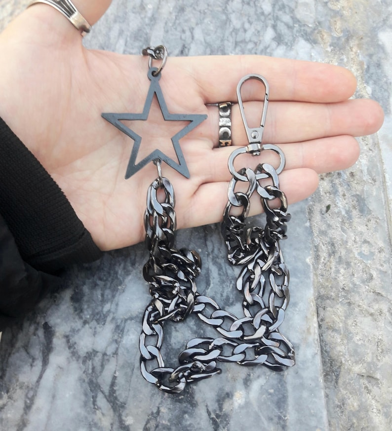 Op de afbeelding: Een zwarte metalen ketting met een stervormige hanger en een karabijnhaak. De ketting is dik en heeft een robuust ontwerp.