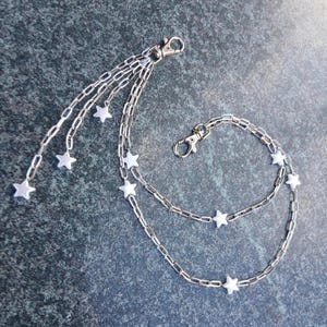 K&ouml;nnte beinhalten: Silbernes Kettenzubeh&ouml;r mit sternf&ouml;rmigen Anh&auml;ngern. Die Kette hat mehrere Str&auml;nge mit Karabinerverschl&uuml;ssen. Das Zubeh&ouml;r ist f&uuml;r die Befestigung an einer Tasche oder als dekoratives Element gedacht. Die Sterne sind wei&szlig;.