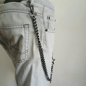 May include: A silver chain with a star charm attached to the end. The chain is hanging from the belt loop of a pair of light gray jeans.