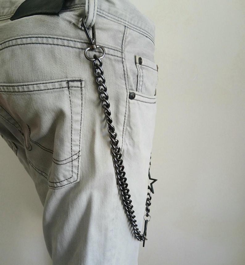 May include: A silver chain with a star charm attached to the end. The chain is hanging from the belt loop of a pair of light gray jeans.