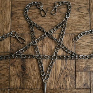 Puede incluir: Una cadena plateada dispuesta en forma de pentagrama, con una larga punta afilada en el punto inferior. La cadena est&aacute; hecha de eslabones entrelazados y tiene tres cierres. El fondo es una superficie de madera.