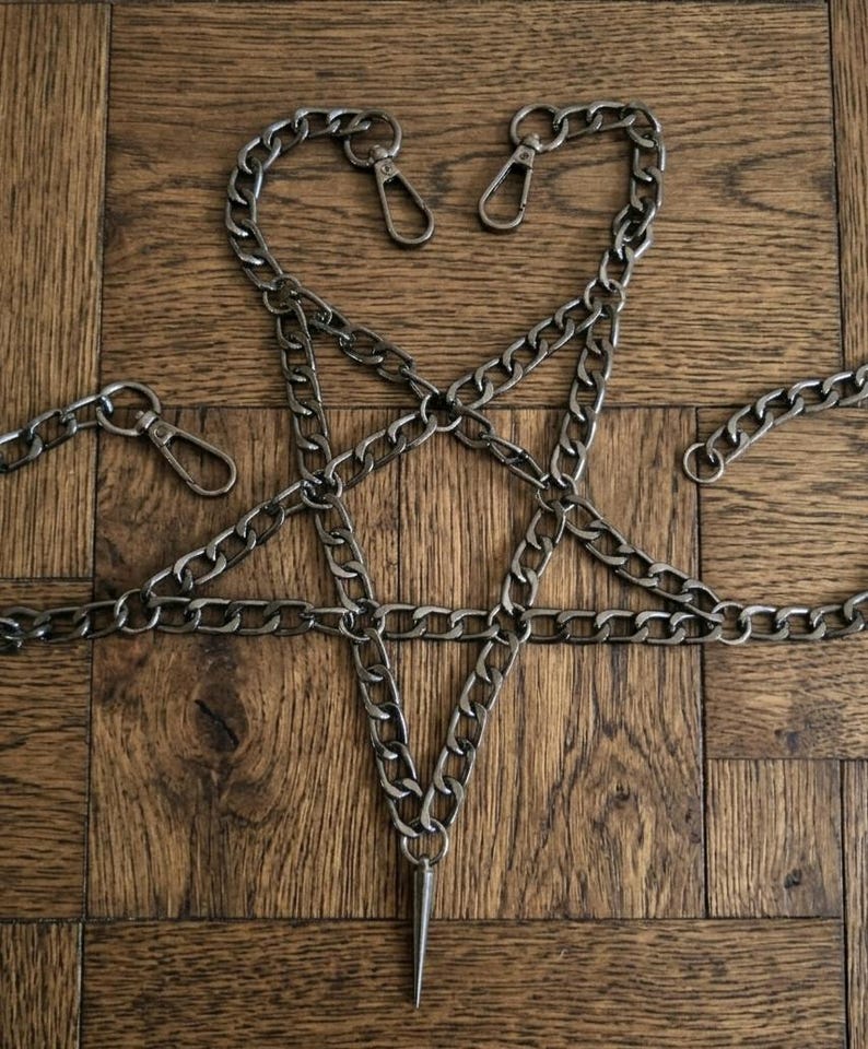 Puede incluir: Una cadena plateada dispuesta en forma de pentagrama, con una larga punta afilada en el punto inferior. La cadena est&aacute; hecha de eslabones entrelazados y tiene tres cierres. El fondo es una superficie de madera.