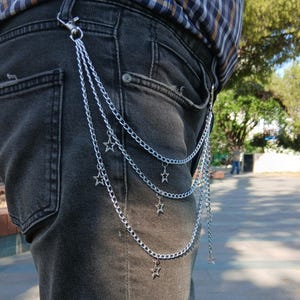May include: Silver chain with star charms attached to a pair of black denim jeans. The chain is attached to the belt loop with a clasp. The chain hangs down in three strands, each with star-shaped charms.