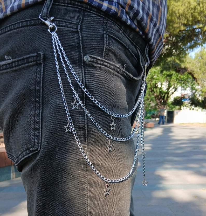 May include: Silver chain with star charms attached to a pair of black denim jeans. The chain is attached to the belt loop with a clasp. The chain hangs down in three strands, each with star-shaped charms.